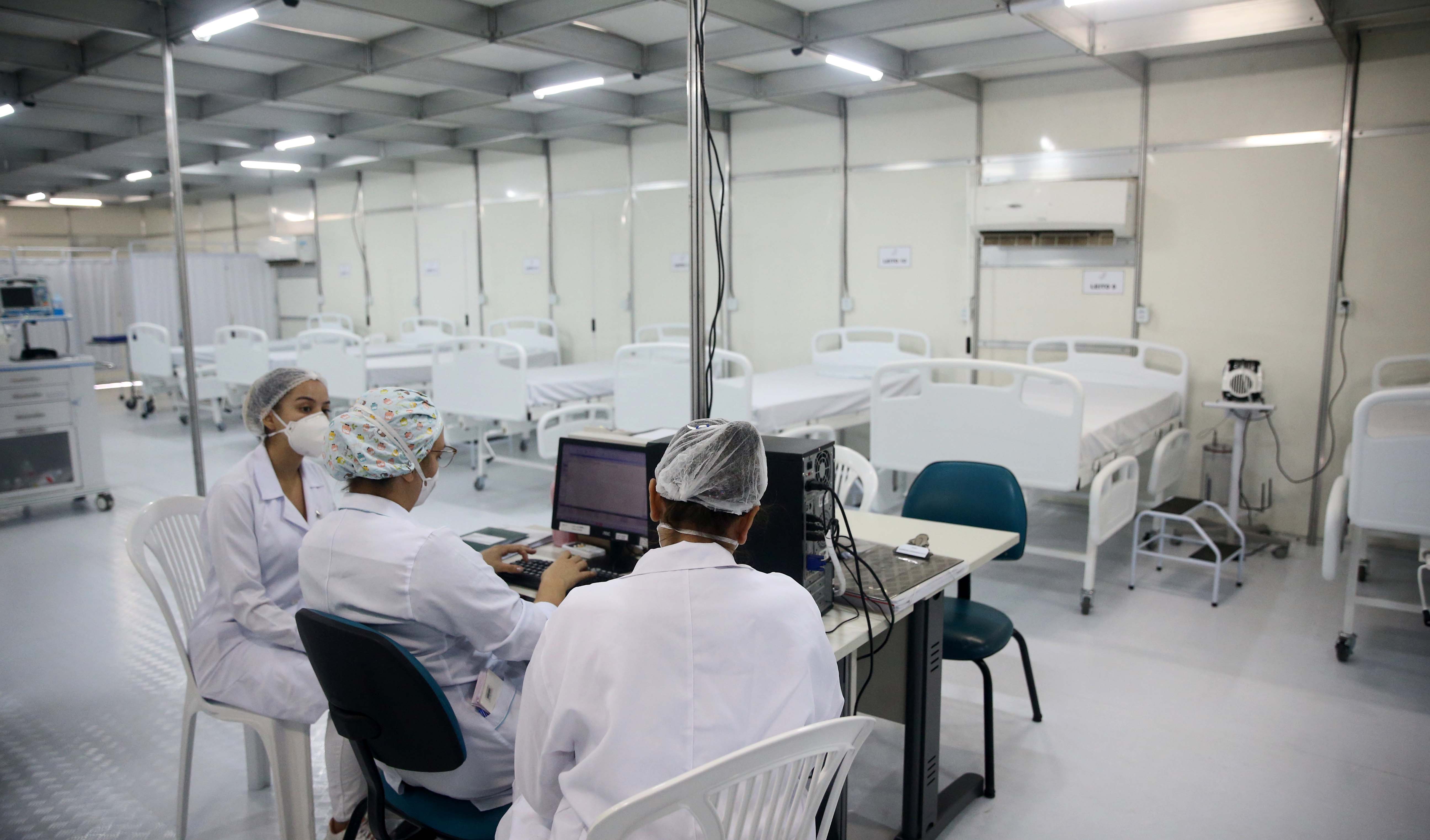 A foto mostra três enfermeiras sentadas diante de uma mesa com um computador, elas estão numa enfermaria da UPA e há quatro leitos ainda vazios na sua frente, pois a ala ainda está em obras